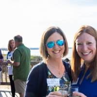 Two women standing outside, smiling at camera, while group of 4 chat behind them